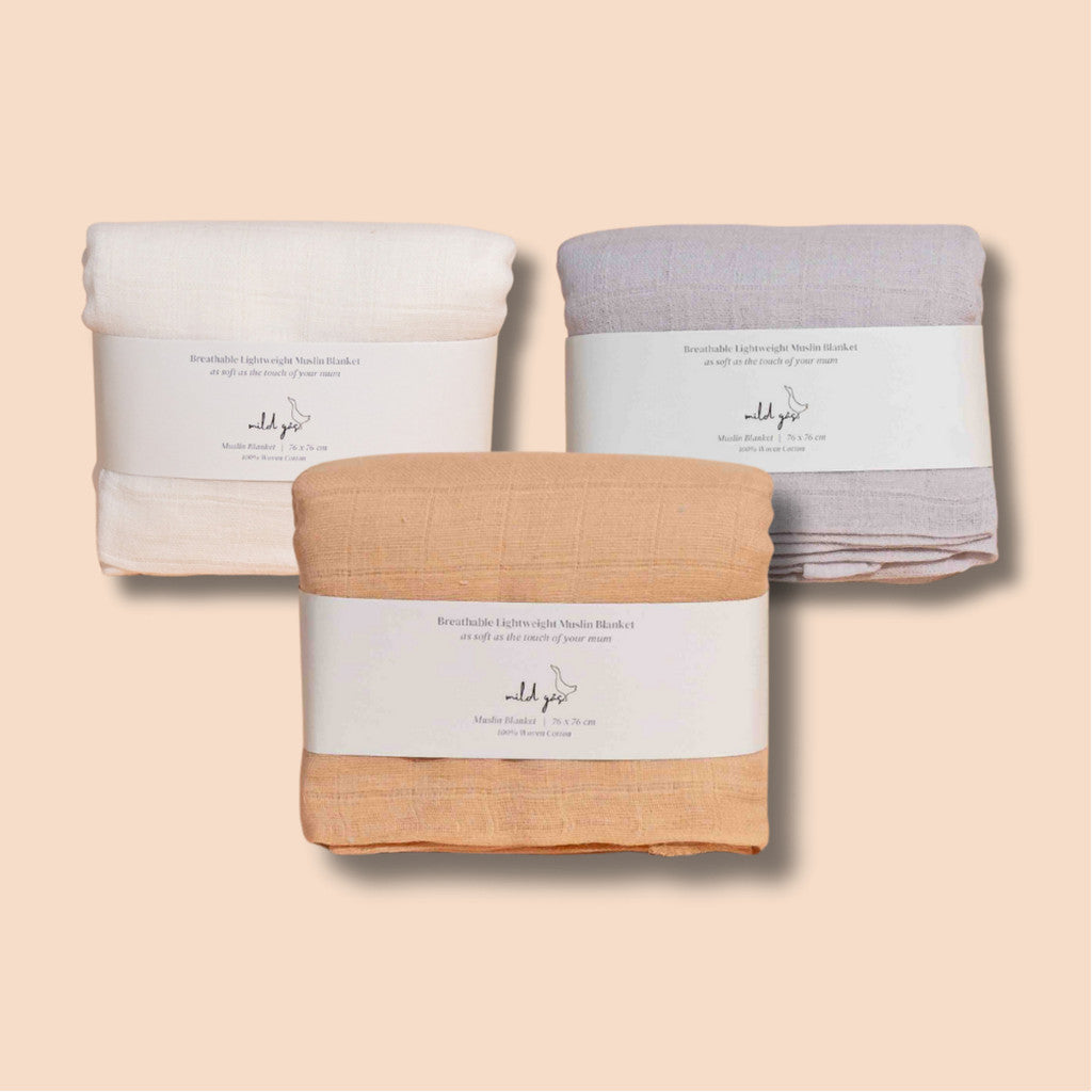 Three folded breathable muslin baby blankets in cream, gray, and peach with minimal labels on a beige background.