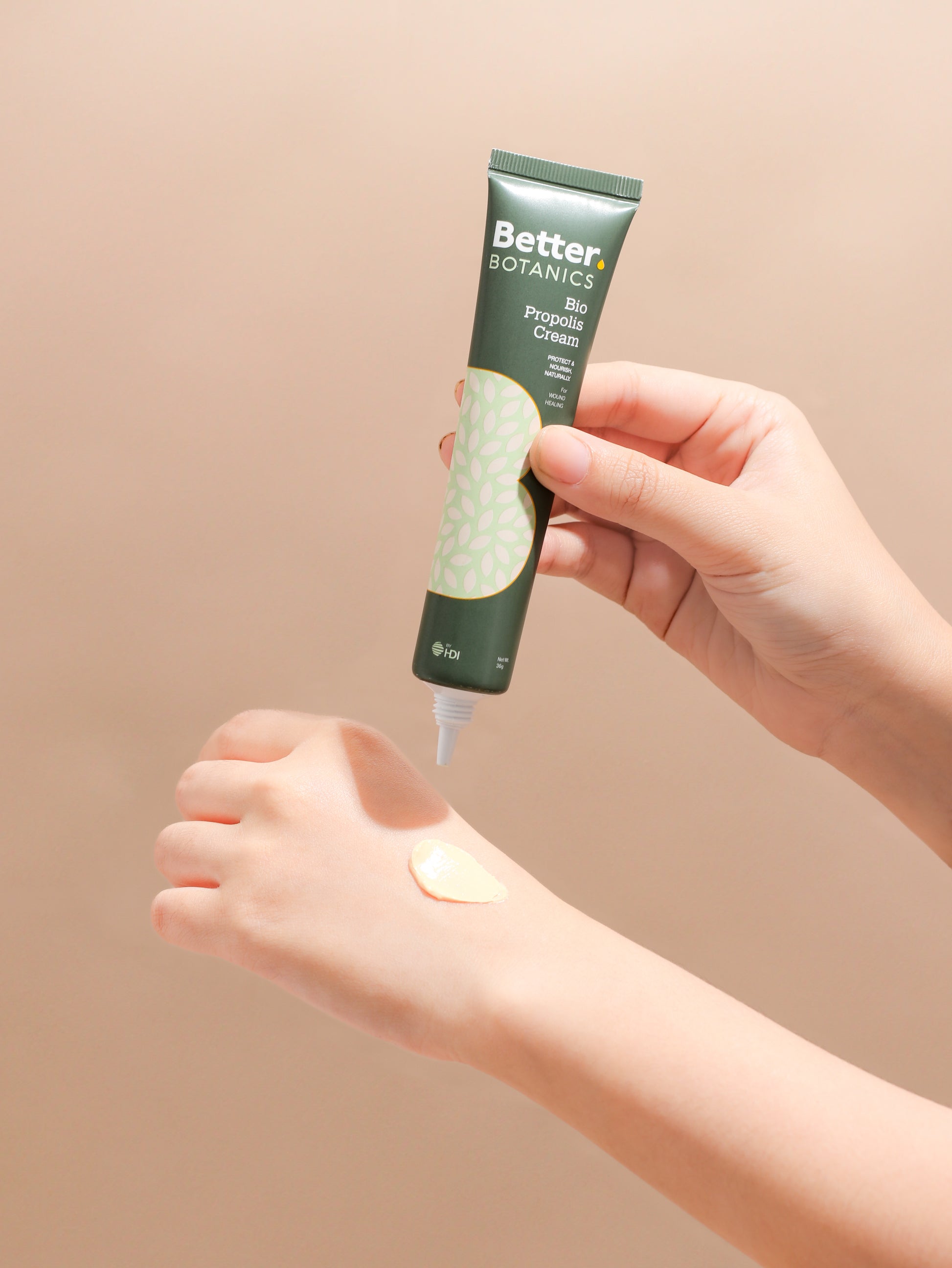 Hand applying a tube of Better Botanics product to a wrist against a beige background