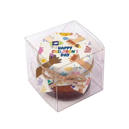 Small honey jar gift in a transparent clear box, wrapped in colorful Happy Children's Day packaging with a twine bow.