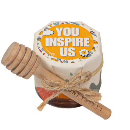 A small glass honey jar with a floral design and a label that reads 'YOU INSPIRE US', accompanied by a honey dipper with a wooden handle tied with a string.