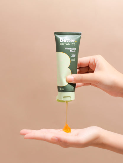 Hand holding a tube of 'Better Botanics' Overnight Mask with product being squeezed out on a beige background