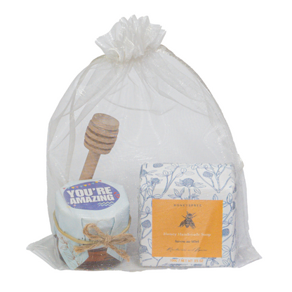 A gift bag containing a honey jar with a honey dipper, a handmade soap with packaging, and a double clear box with a floral design. All items are wrapped in a transparent organza bag.