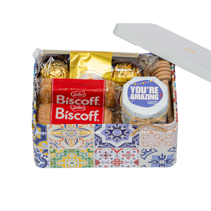 A tin with a colorful patterned design, open to reveal an assortment of sweet treats including Biscoff cookies, a Ferrero Rocher, and a honey dipper.