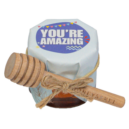 Small multi-floral honey jar with blue lid labeled “You’re Amazing,” with honey dipper tied with string.